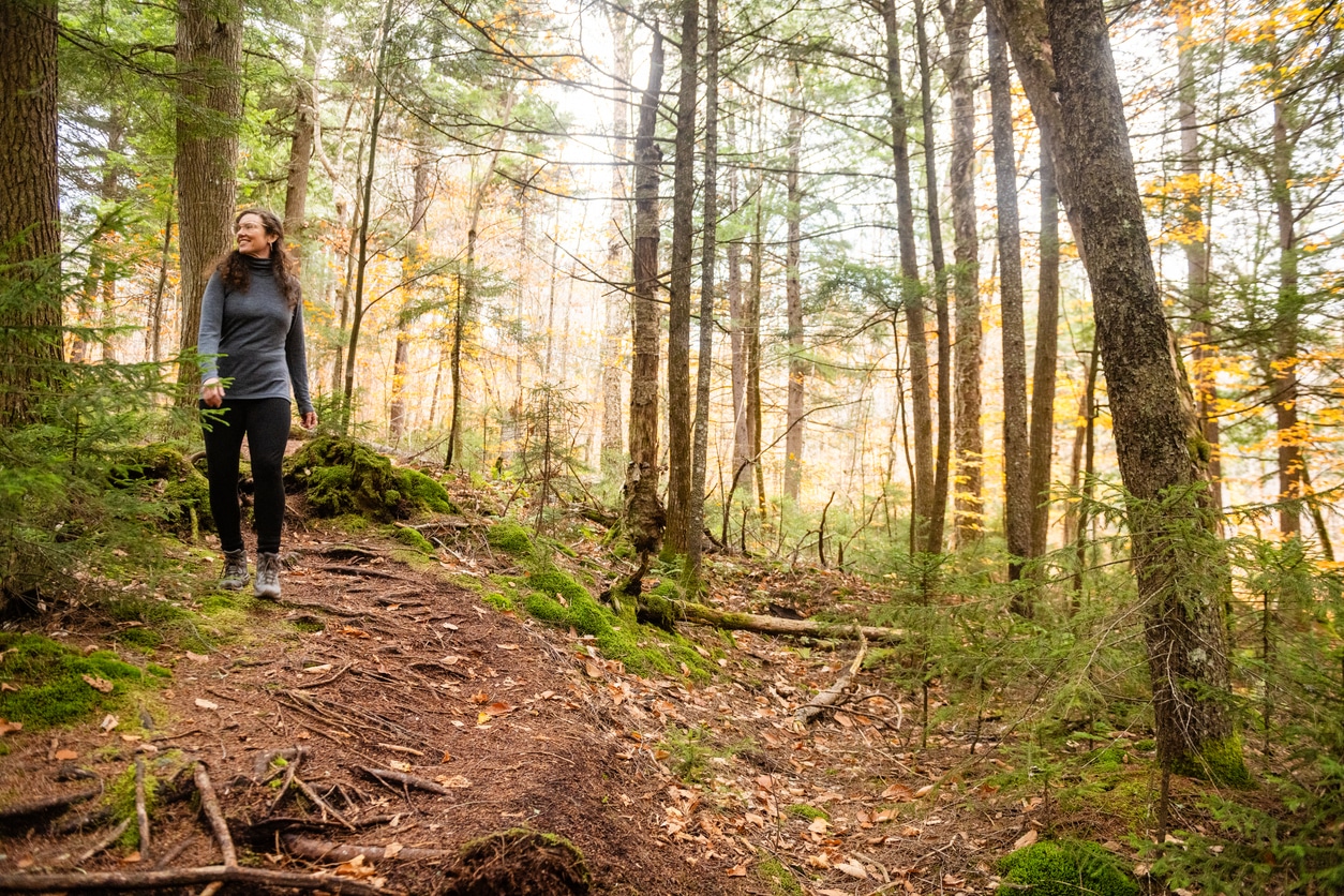This is a photograph of a woman in her 40s wearing long sleeves and pants while hiking on a forest trail in the Adirondack Mountains in New York on an October day.