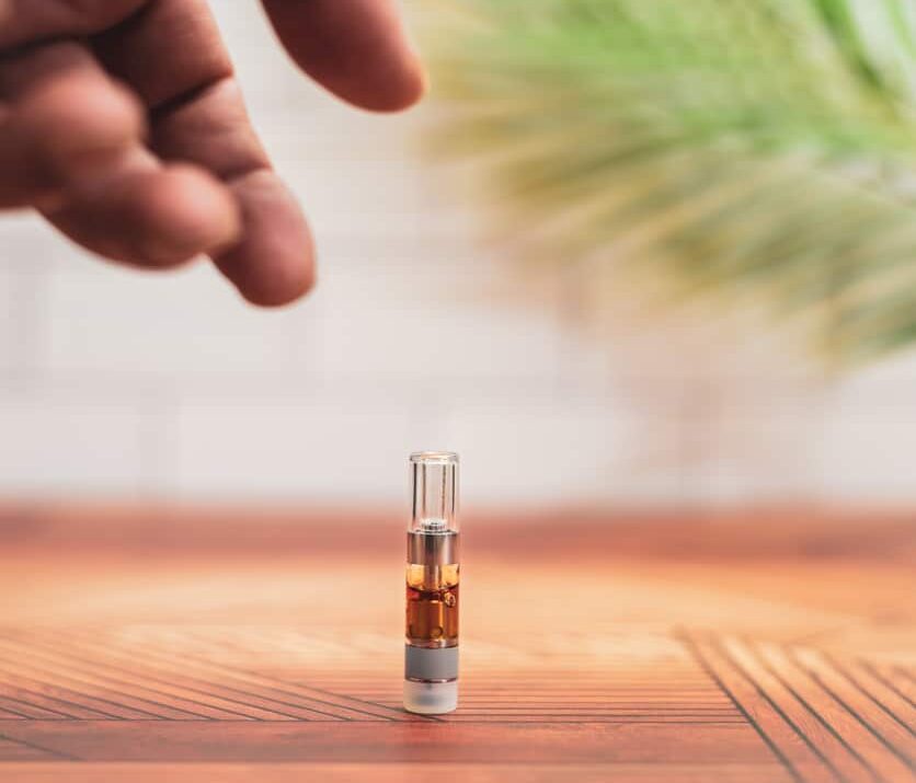 A hand reaches to pick up a cannabis vaporizer cartridge from a counter at home.