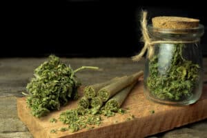 Pre-Roll cannabis joints are placed on the wooden chopping board with cannabis buds in a clear glass jar at the black background