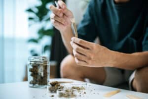 Selective focus and close up hands of young man preparing to smoking marijuana joint Cannabis in living room at home, he rolling a cannabis on paper, dry weed blunt in bottle on table