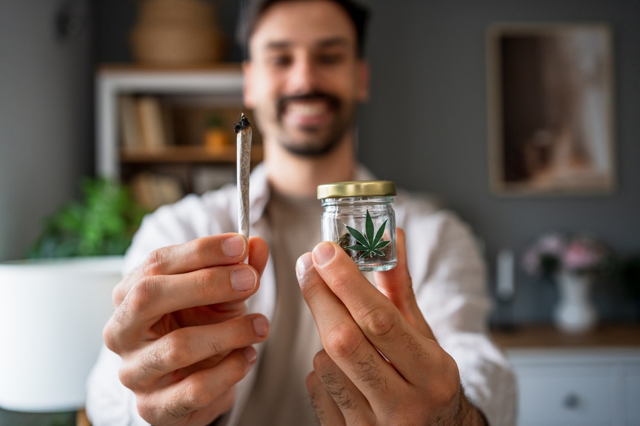 Young adult man holding a marijuana joint while displaying medical cannabis in a jar, enjoying a relaxed moment at home